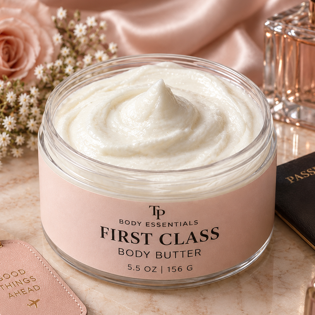 A jar of TP Body Essentials First Class Body Butter filled with white cream, styled on a marble surface with a pink rose, dried flowers, a perfume bottle, and a leather luggage tag.